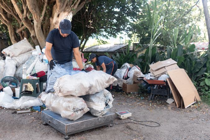 An adult Hispanic man weighs a pile of sacks of reclaimed materials, supported by a woman in the background