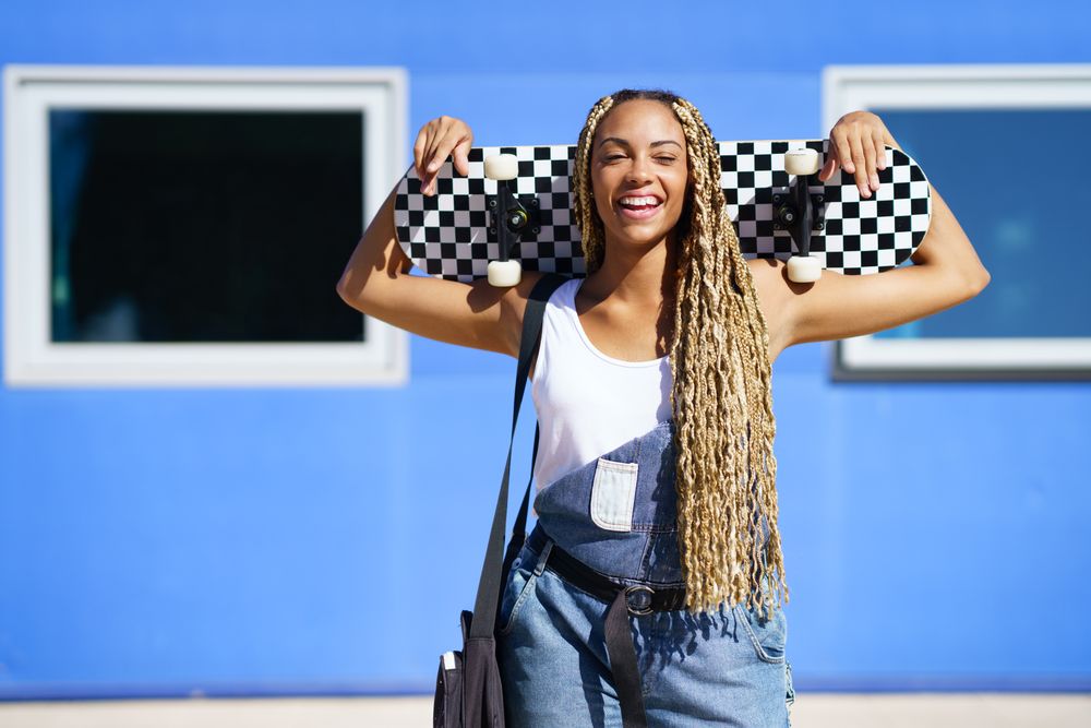 Joyful female in denim overalls standing with skateboard behind head in front of blue building