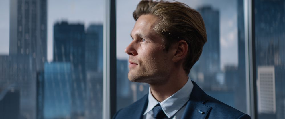 Portrait of Successful Businessman in a Tailored Suit Standing in Office Looking out of the Window on Big City with Skyscrapers.