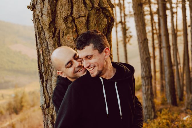 Smiling men hugging near tree in forest on hill