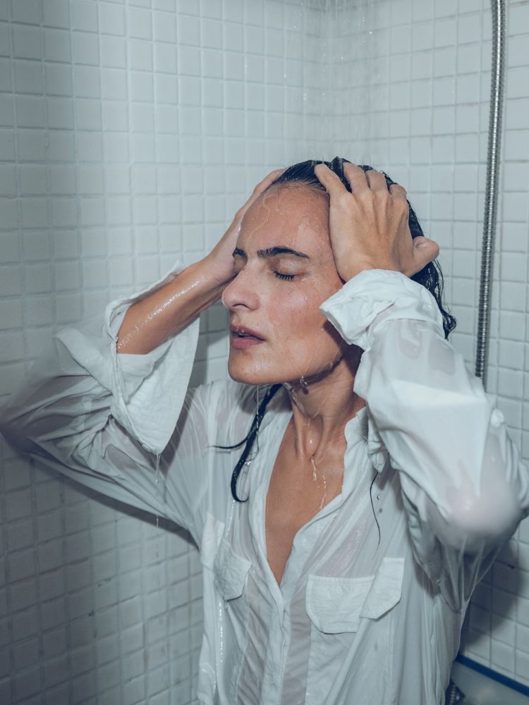 Upset woman touching head while taking shower in wet shirt