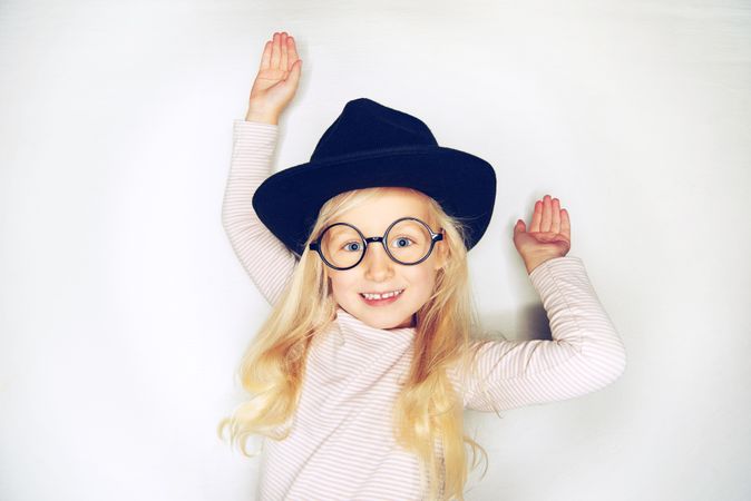 Smiling blonde girl with pursed lips wearing hat and glasses with her arms up