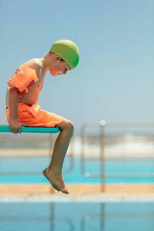 Cute boy in swim gear sitting on edge of spring board