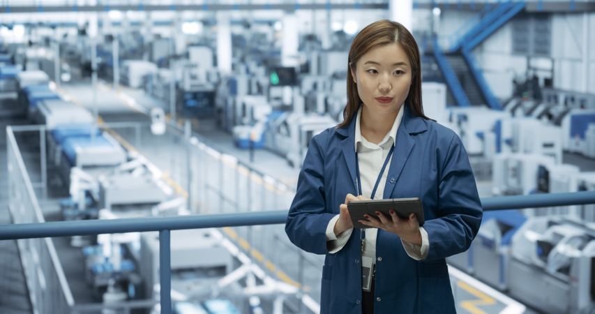 Thoughtful Asian Engineer Monitoring and Analyzing Conditions at a Pristine Modern Electronics Factory with Automated Robot Hands Working with the Help of Artificial Intelligence Software