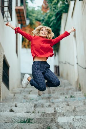 Smiling blonde woman with red shirt jumping on steps outdoors