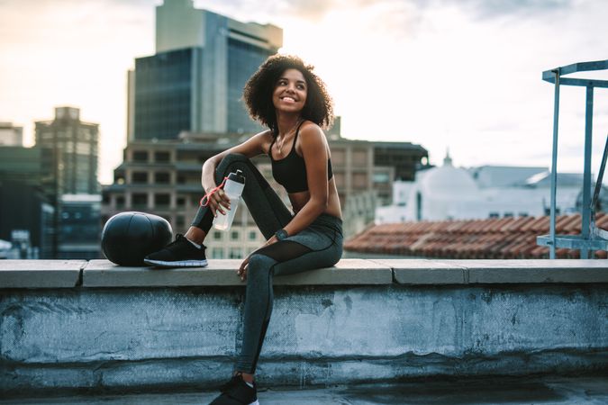 Smiling fitness woman sitting on terrace of a building drinking water