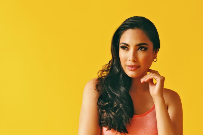 Headshot of calm Hispanic woman looking away in yellow room, with hand up to her cheek
