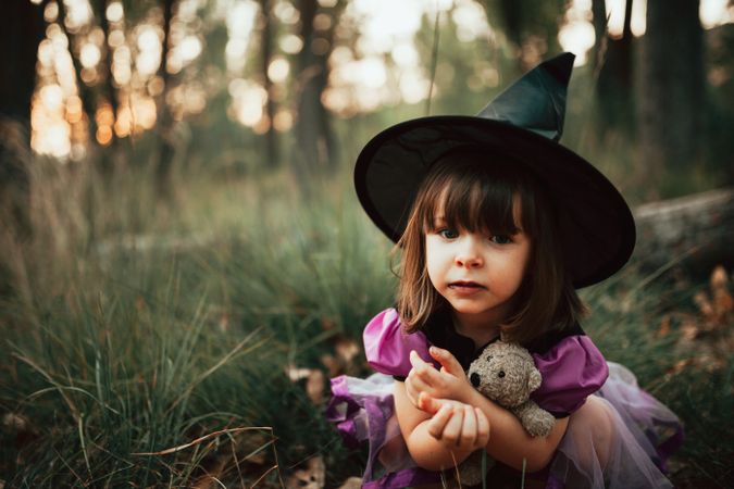 Somber girl in witch costume crouched down in the forest with her teddy bear