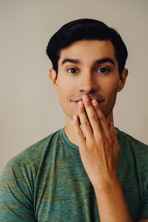 Portrait of Hispanic male in neutral room with hand over his mouth, vertical