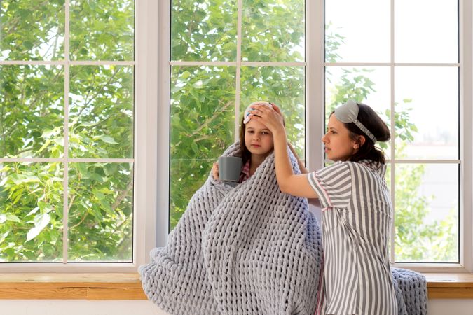 Mother caring for sick daughter at home near window.
