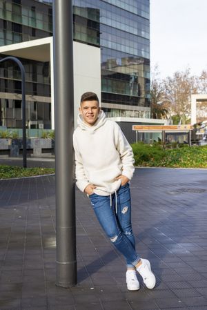 Portrait of young adult with hands in pocket leaning on pole outside, vertical