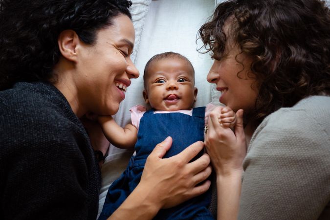 Two smiling moms and a baby with her tongue out while lying on a bed