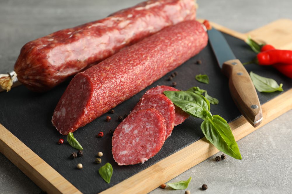 Two sticks of cured meats with slices on wooden board with knife