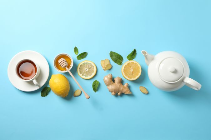 Flat lay. Cup of tea, teapot, lemon, ginger, mint, honey and dipper on blue background, copy space