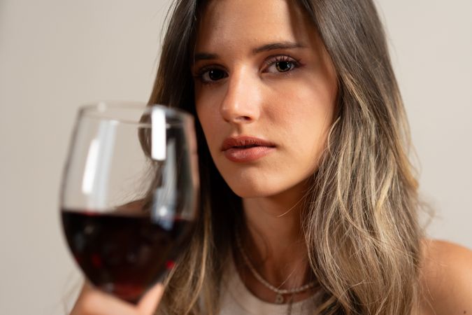 Elegant woman holding glass of red wine, thoughtful expression in dim light