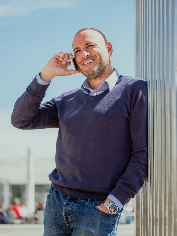 vertical photo of a businessman leaning on a wall of a building and talking to the mobile smiling