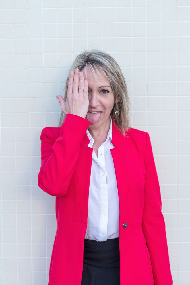 Portrait of a blonde woman wearing red jacket against a tiled wall covering her eye with tongue out