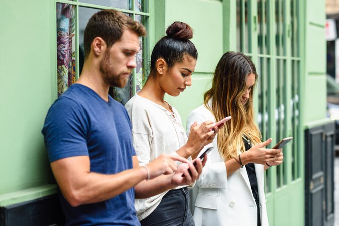 Two female and one male friend looking down at their phones leaning on green wall