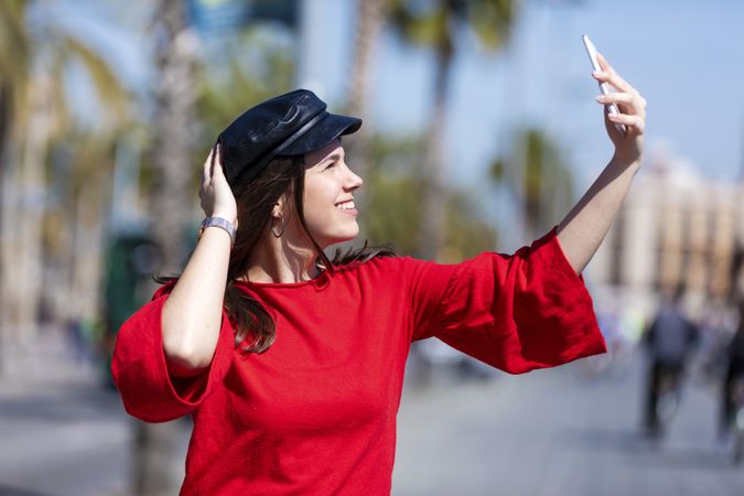 Side view of woman wearing red blouse sitting on a metallic fence while taking a selfie outdoors in the street on a bright day