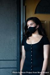 Portrait of woman looking out of her doorway wearing PPE mask and dress 5wXxA4