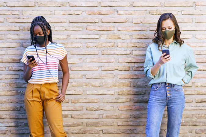 Female friends in face masks texting on phone in front of brick wall