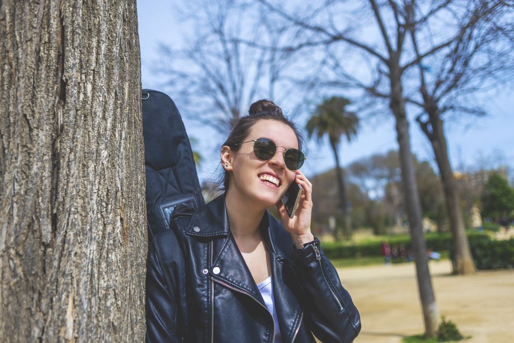 Side view of a young laughing hipster woman standing and leaning on a tree at park in sunny day while using a mobile phone