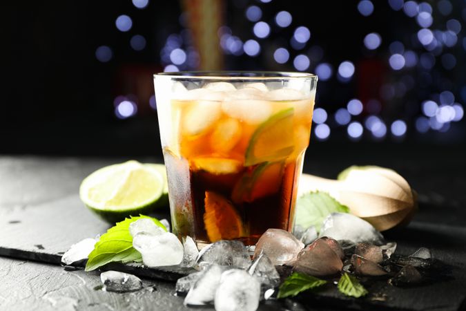 Glass with cola and lime slices, juicer, ice, mint against blurred lights background