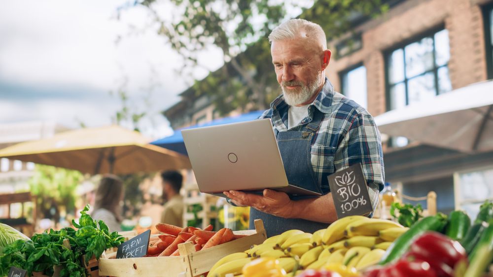 Successful Farmer Working on a Laptop Computer, Writing Emails to Suppliers to Orders Produce for His Organic Street Vendor Stall with Sustainable Fruits and Vegetables