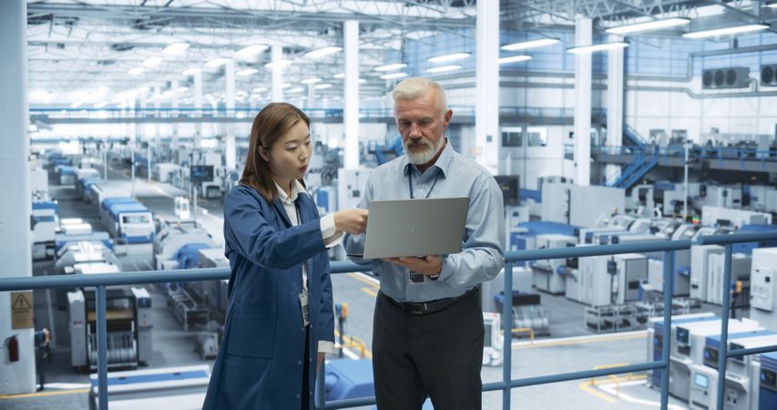 Male Engineer and Female Project Manager Standing on a Platform, Talking, Using Laptop Computer and Monitoring Production at a Modern Automated Electronics Manufacture with AI Robots