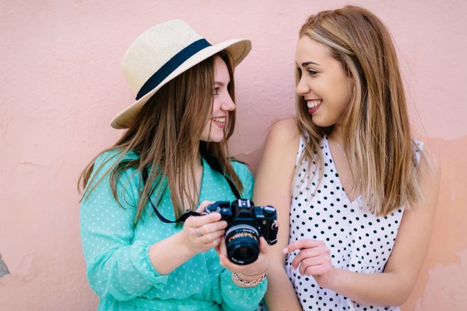 Two smiling women holding a camera and standing against pink wall