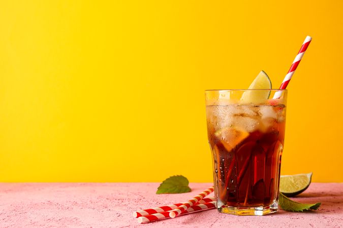 Glass with cold cola, tubule and lime slice against yellow background