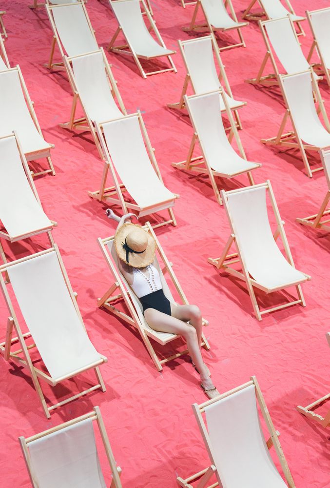 Woman in bathing suit wearing a sun hat lying on one of the arranged beach lounger on pink surface