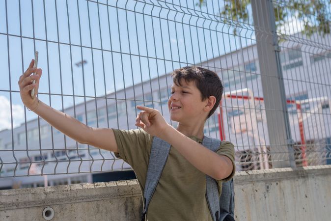 Smiling boy pointing while taking selfie on smartphone outside of school yard