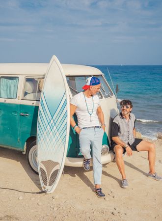Men with surfboard near car on dusty seaside