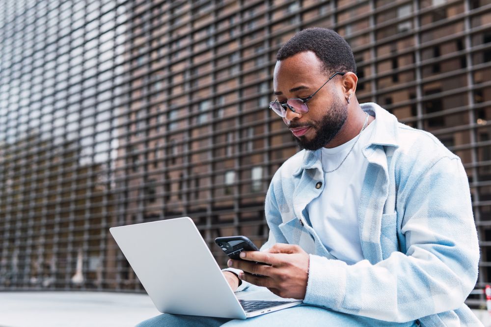 Black man browsing mobile phone and laptop on street