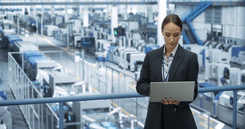 Young Female Engineer Standing on a Platform, Using Laptop Computer at an Electronics Factory. Machines are Undergoing Maintenance, Technician Monitoring Updates Through Online Software