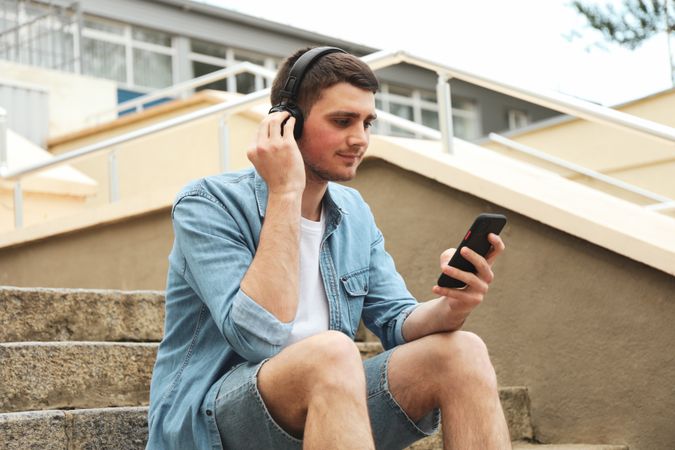 Young man in headphones sits on the steps and looking in phone