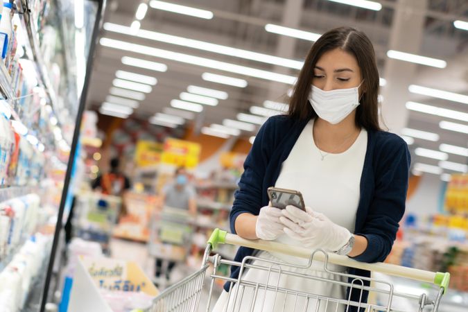 Woman checking phone over grocery cart wearing surgical mask