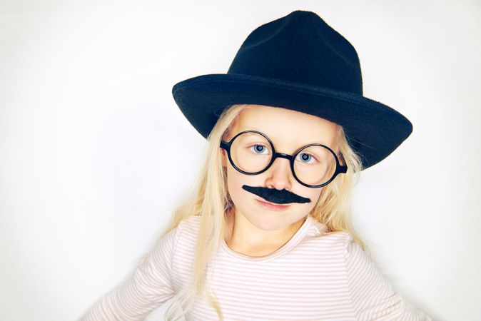 Unsmiling blonde girl wearing fake mustache, hat and glasses