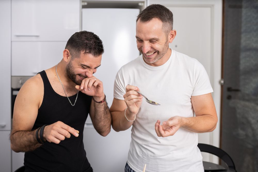 Laughing male couple tasting pasta as they prepare meal