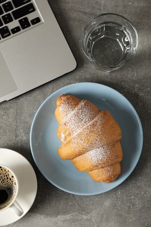 Vertical composition, top view of plate with croissant with water glass and laptop