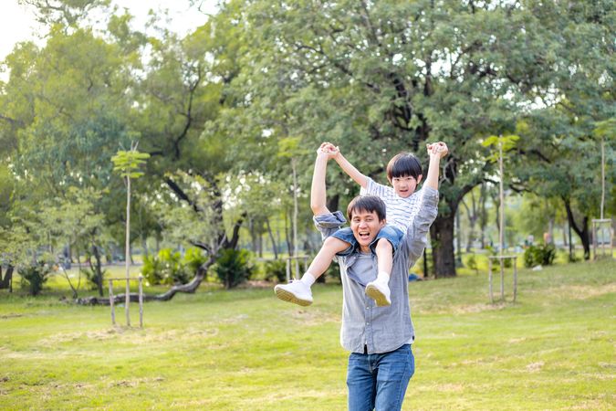 Asian playful boy sitting on father shoulder playing at park in summer