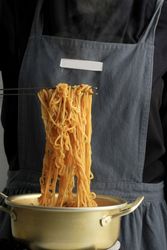 Cook holding up noodles with chopsticks cooking in brass pot