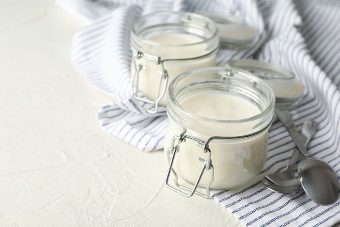 Glass jars with sour cream yogurt, spoons and towel on cement background