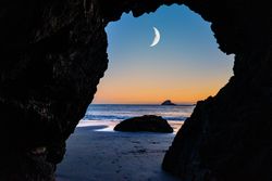 Crescent moon seen between rocky arch on quiet beach at dusk