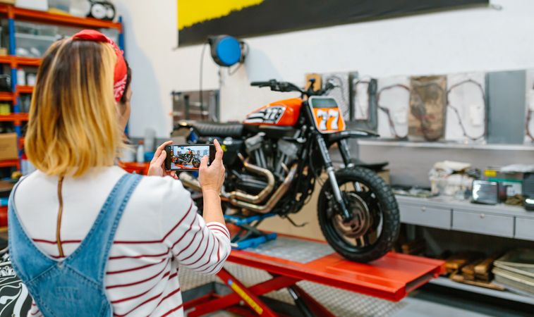 Female mechanic taking photo to motorcycle repaired over platform on garage