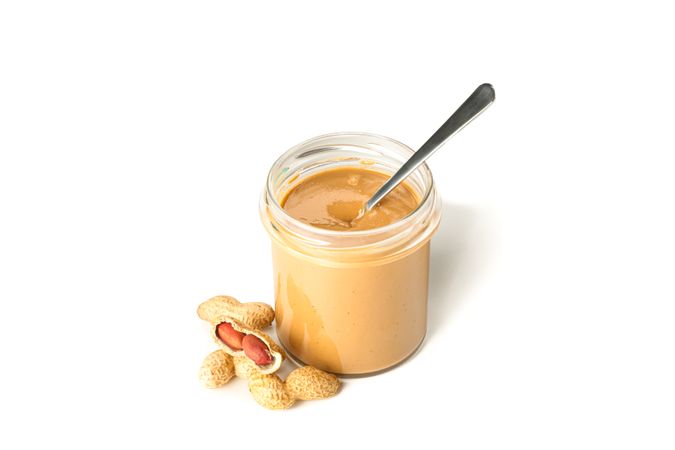 Creamy peanut butter in glass jar, peanut and spoon isolated on light background. A traditional product of American cuisine