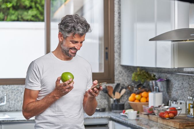 Smiling mature man with apple and smartphone in kitchen