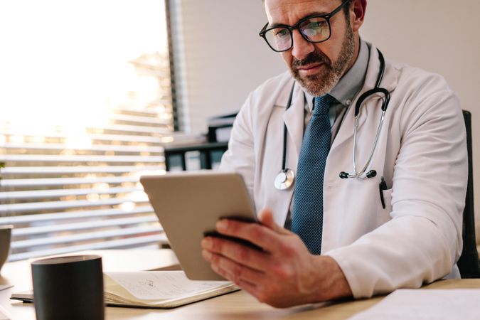 Male doctor looking at digital tablet while making notes in his office