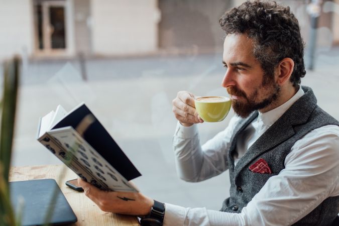 Content man with curly hair drinking coffee in cafe while reading book